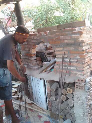 The image depicts a man constructing a brick oven, standing beside it with tools and materials scattered around, set against a backdrop of trees and foliage.