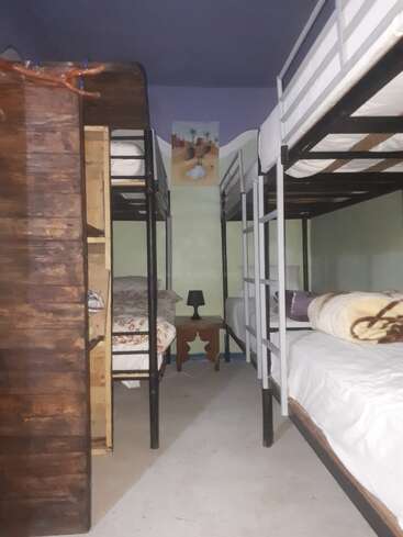 The image depicts a room with bunk beds, featuring wooden and metal frames, and a small table with a lamp, set against a light green wall and purple ceiling.