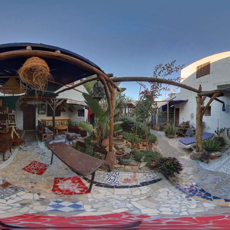 The image depicts a serene courtyard with a mosaic floor, featuring a pergola, lounge chairs, and lush greenery, set against a clear blue sky.