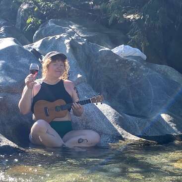 Uma mulher está sentada em uma pedra em águas rasas, tocando um ukulele e segurando uma taça de vinho tinto, vestindo uma camiseta preta e calça verde.