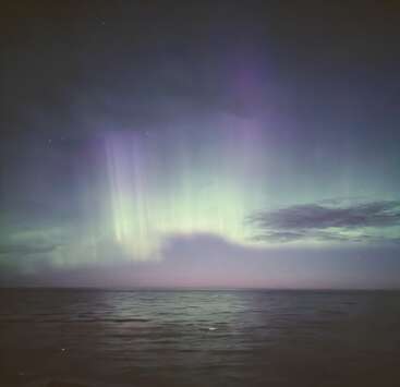 Ondas suaves ondulam em um mar escuro, enquanto as etéreas luzes verdes e roxas do norte dançam acima, iluminando o céu noturno com um brilho mágico e sereno.
