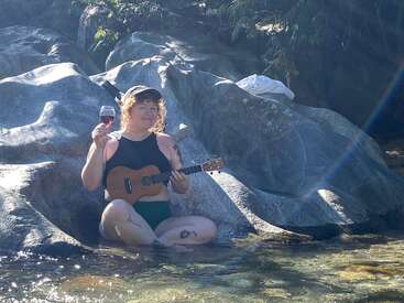 Uma mulher está sentada em uma pedra em águas rasas, tocando um ukulele e segurando uma taça de vinho tinto, vestindo uma camiseta preta e calça verde.