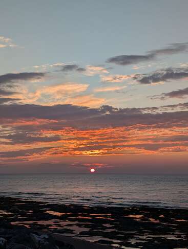 Um pôr do sol deslumbrante sobre o oceano, com nuvens laranja e rosa vibrantes, ondas suaves, uma costa rochosa e o sol baixo no horizonte. Atmosfera tranquila.