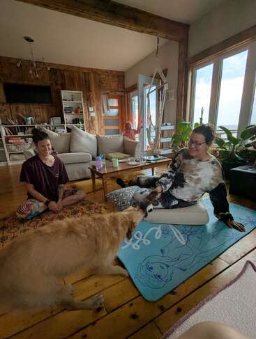 Duas pessoas relaxam em uma aconchegante sala de estar de madeira. Um cachorro grande brinca de cabo de guerra com uma delas. Uma criança fica ao lado da porta aberta.