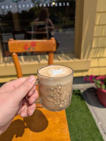 Uma mão segura uma xícara de vidro de café com arte latte. A luz do sol incide sobre uma mesa e uma cadeira de madeira, com flores e uma janela de café ao fundo.