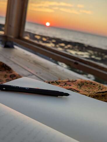 Uma caneta preta repousa sobre um caderno aberto ao lado de uma janela, com vista para um oceano sereno ao pôr do sol, convidando à reflexão pacífica e à inspiração criativa em uma luz quente.
