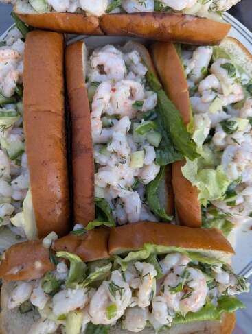 Esta imagem mostra vários rolinhos de lagosta recheados com uma salada cremosa de camarão, aipo picado, ervas e alface, servidos em pães torrados, dispostos bem próximos uns dos outros.