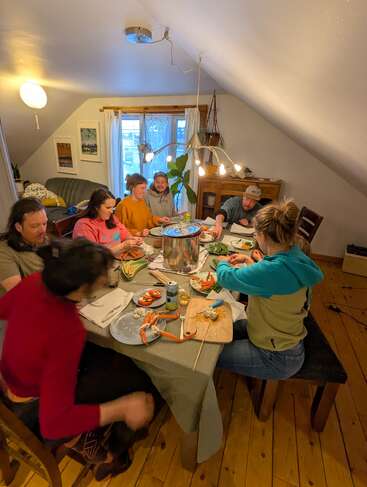 Sete pessoas se reúnem em torno de uma mesa de jantar, compartilhando comida e conversando em uma sala aconchegante e bem iluminada, com piso de madeira e decoração caseira, evocando conforto e união.