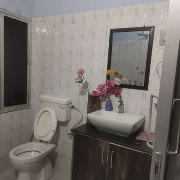 This bathroom features a white toilet, a rectangular sink on a dark cabinet, a wall mirror, floral decorations, soap dispenser, tiled walls, and a frosted window.