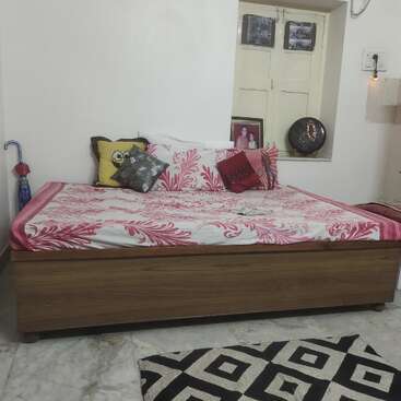 A cozy room with a wooden bed covered in a pink floral bedsheet, colorful cushions, an umbrella, framed photos on a windowsill, and a geometric rug.