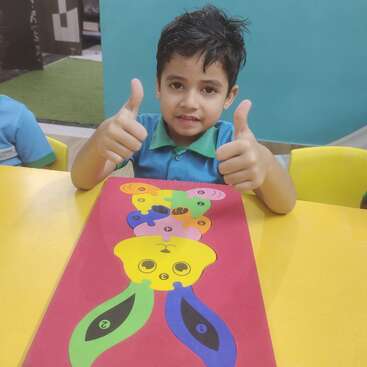 A cheerful boy sits at a yellow table, giving two thumbs up, proud of his colorful paper craft project featuring animal shapes on a red sheet.