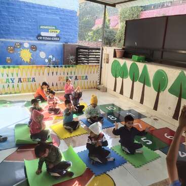 Em uma área de recreação interna cheia de cores, as crianças pequenas sentam em tapetes, participando de uma atividade em grupo. A luz do sol brilha na sala, criando um clima alegre e propício para o aprendizado.