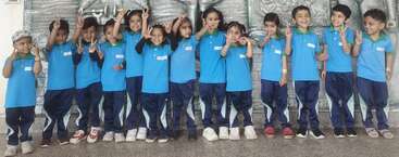 Um grupo de crianças pequenas, com uniformes azuis iguais, sorri alegremente, fazendo sinais de paz, em pé juntas em frente a um fundo prateado texturizado, irradiando alegria.