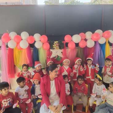Um grupo de crianças pequenas e uma professora comemoram o Natal, vestidos com roupas festivas e chapéus de Papai Noel, rodeados por balões, decorações coloridas e sorrisos felizes.