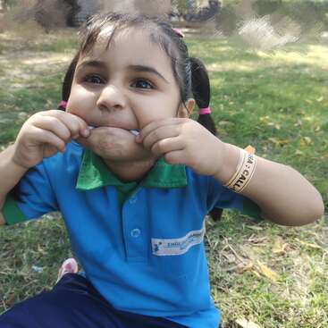 A young girl in a blue uniform sits on grass, playfully stretching her mouth with her fingers, making a funny face. She looks cheerful and lively.