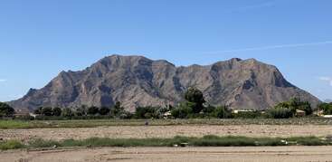 Ein schroffer Berg steht unter einem klaren blauen Himmel. An seinem Fuße stehen grüne Bäume und kleine Häuser, im Vordergrund sind sandiger Boden und niedriges Gras zu sehen.