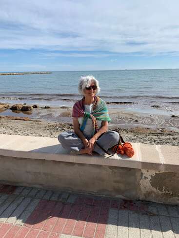 Das Bild zeigt eine ältere Frau, die auf einer Steinbank am Meer sitzt, mit einer ruhigen Strandszene und einem blauen Himmel mit Wolken im Hintergrund.