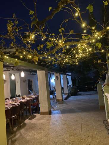 L'image représente une salle à manger extérieure sereine de nuit, avec une pergola ornée de guirlandes lumineuses, une table dressée pour le dîner et une voiture garée à l'arrière-plan.