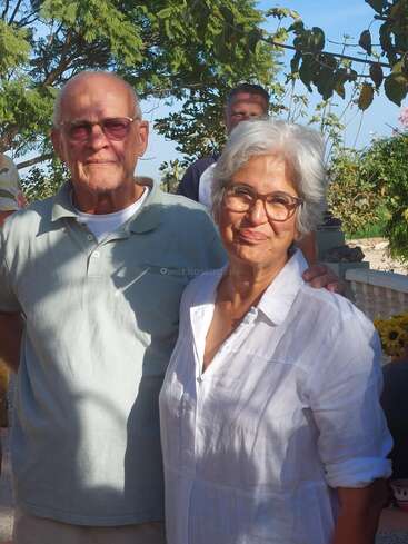Das Bild zeigt ein älteres Paar, das zusammen steht. Der Mann auf der linken und die Frau auf der rechten Seite tragen beide weiße Hemden und sind von Bäumen und einem blauen Himmel umgeben.