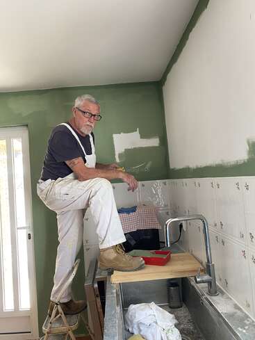 An older man stands on a chair in a kitchen, painting the wall green. He wears glasses, overalls, and boots, working carefully near the ceiling.