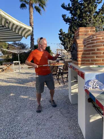 A man in an orange shirt is making pizza outdoors, using a brick oven. Palm trees, chairs, and a striped canopy create a relaxed, sunny atmosphere.