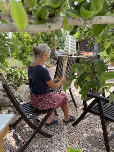 A woman with gray hair sits outdoors on a dark wooden chair, crocheting or knitting. She’s surrounded by green foliage, gravel, and another person in the background.