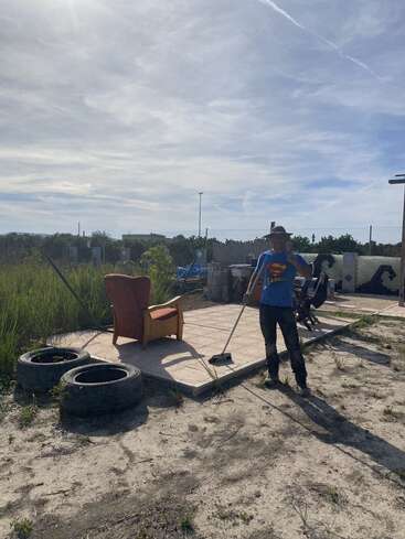 A person wearing a hat and Superman shirt sweeps a tiled outdoor area. An armchair, tires, and scattered grass complete this bright, partly cloudy scene.