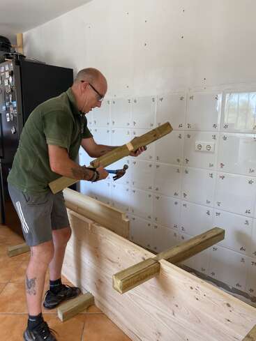 A man wearing glasses and casual clothes is building a wooden structure indoors, using a hammer. He is focused on assembling beams in a tiled room.
