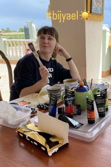 A woman sits at a table outdoors, holding a paintbrush, surrounded by paints, art supplies, and a teapot. The hashtag #bijyael☀️ appears above her.