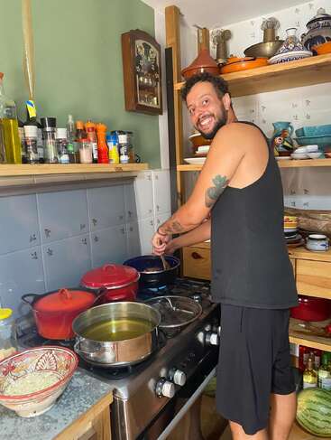 A smiling man in a black tank top cooks at a cozy kitchen stove, surrounded by pots, spices, colorful dishes, and shelves filled with various kitchenware.