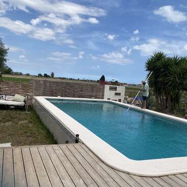 A imagem mostra uma cena serena no quintal, com uma piscina, um deck de madeira e um homem de pé na borda da piscina, segurando um skimmer.
