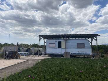 Ein weißer Wohnwagen steht unter einem Holzunterstand in einer grasbewachsenen, ländlichen Gegend. Im Vordergrund blühen Wildblumen, über ihnen ist der Himmel teilweise bewölkt. Friedliche Atmosphäre.