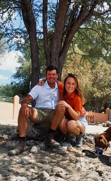 A smiling man and woman pose together outdoors under a large tree, sitting on rocks. A goat drinks from a can near them. Sunny day. Relaxed atmosphere.