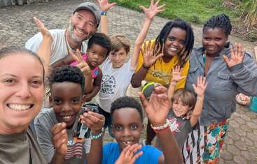 Eine fröhliche Gruppe von Erwachsenen und Kindern lächelt und winkt begeistert in die Kamera. Sie stehen im Freien auf einer gepflasterten Fläche und strahlen Glück und Zusammengehörigkeit aus.