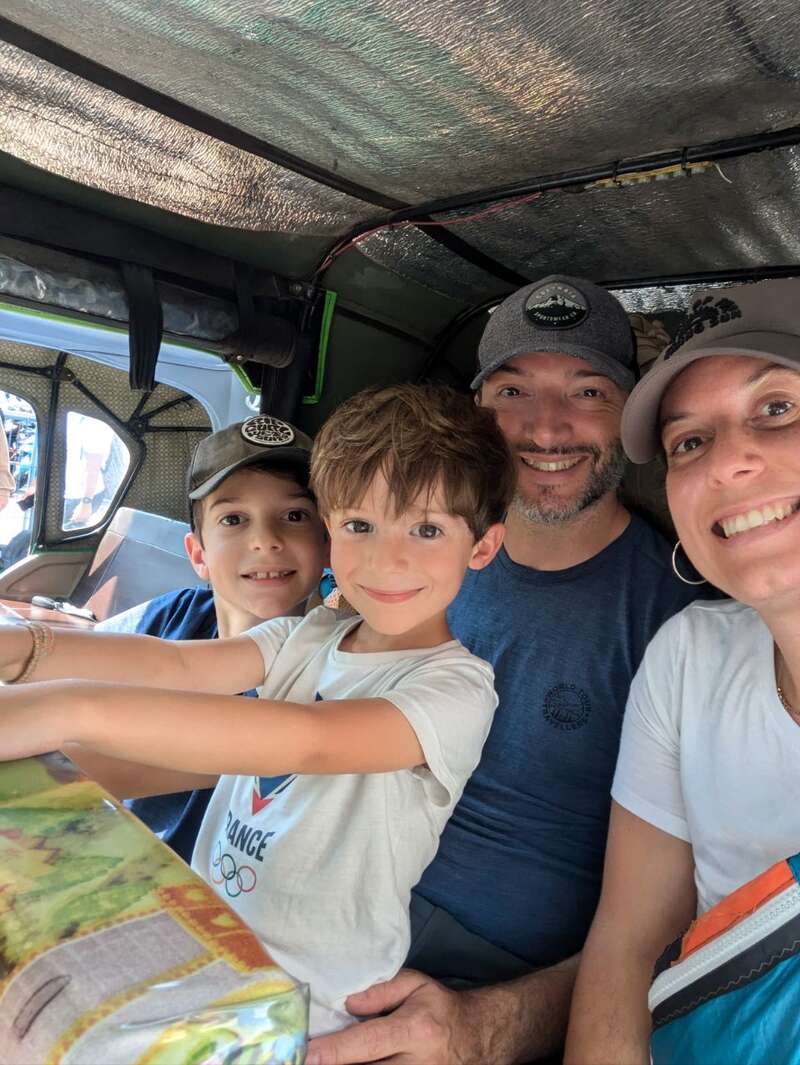 Eine lächelnde vierköpfige Familie, darunter zwei kleine Jungen, sitzt eng beieinander in einem Fahrzeug, vielleicht auf einer lustigen Fahrt oder einem Reiseabenteuer, und genießt den Moment.
