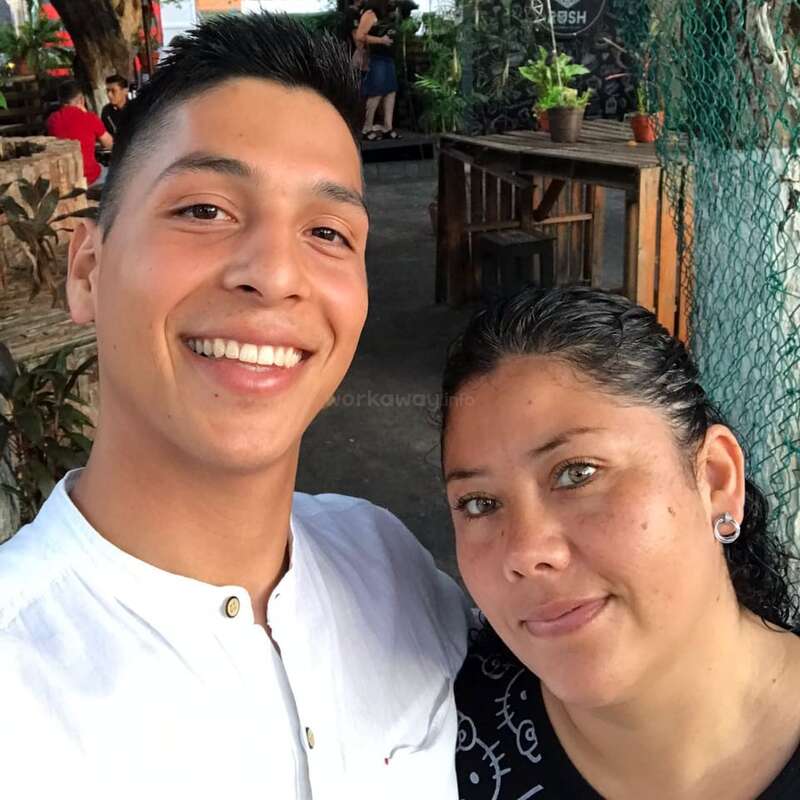 Ein junger Mann und eine Frau lächeln für ein Selfie im Freien. Hinter ihnen stehen Holztische, Grünpflanzen, Menschen und eine entspannte, lockere Atmosphäre.