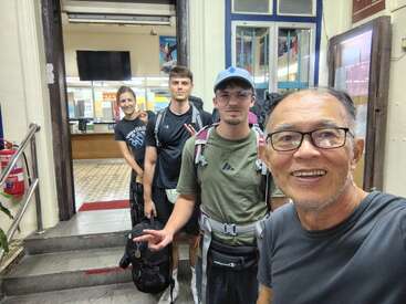 Four people, likely travelers with backpacks, pose for a selfie at what appears to be a station or building entrance, smiling and ready for adventure.