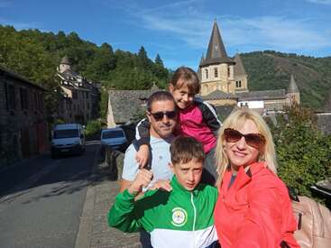 Une famille souriante de quatre personnes pose pour un selfie en plein air, avec en arrière-plan des bâtiments historiques en pierre, une église avec un haut clocher et des collines verdoyantes.