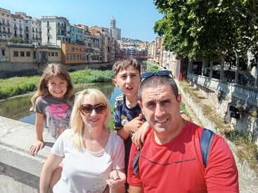 Une famille de quatre personnes sourit pour une photo lors d'une journée ensoleillée, debout sur un pont enjambant une rivière, avec des bâtiments et des arbres colorés derrière eux.