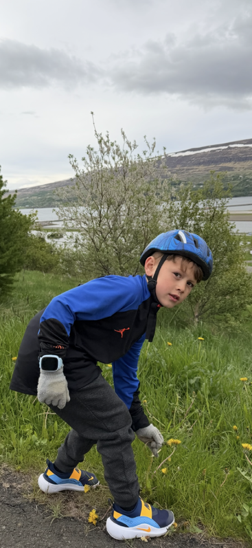 Ein kleiner Junge mit blauem Helm und Handschuhen hockt am grasbewachsenen Straßenrand, im malerischen Hintergrund sind Hügel, Bäume und ein Fluss zu sehen.