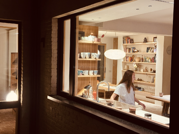 A woman is seen through a window in a cozy, warmly lit kitchen filled with shelves, books, cups, and decorations, giving a homely atmosphere.