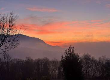 A vibrant sunset bathes the landscape in warm orange and pink hues. Silhouetted trees, mist, and distant hills create a peaceful, atmospheric evening scene.