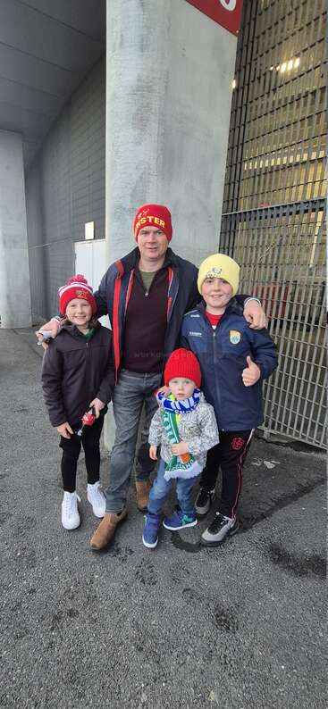 Um homem e três crianças, todos usando chapéus coloridos, estão sorrindo do lado de fora de um estádio. As crianças parecem felizes e uma delas faz sinal de positivo com o polegar.