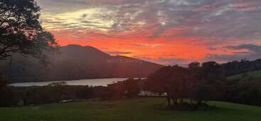 Um pôr do sol de tirar o fôlego ilumina o céu com tons vibrantes de laranja e rosa sobre um lago sereno, colinas em silhueta e campos verdejantes pontilhados de árvores.