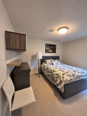 Un acogedor dormitorio con una cama de matrimonio, ropa de cama con motivos de hojas, una silla blanca, armarios de madera, una estantería suspendida, una lámpara de pie, una alfombra y un cuadro con motivos naturales.