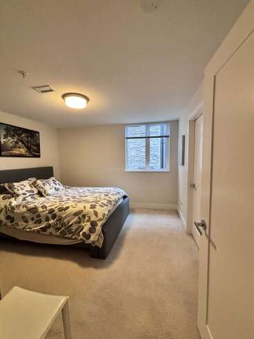 Se trata de un dormitorio acogedor y minimalista con moqueta beige, una cama de matrimonio con ropa de cama estampada, una ventana con persianas, paredes blancas y unos sencillos cuadros en las paredes.