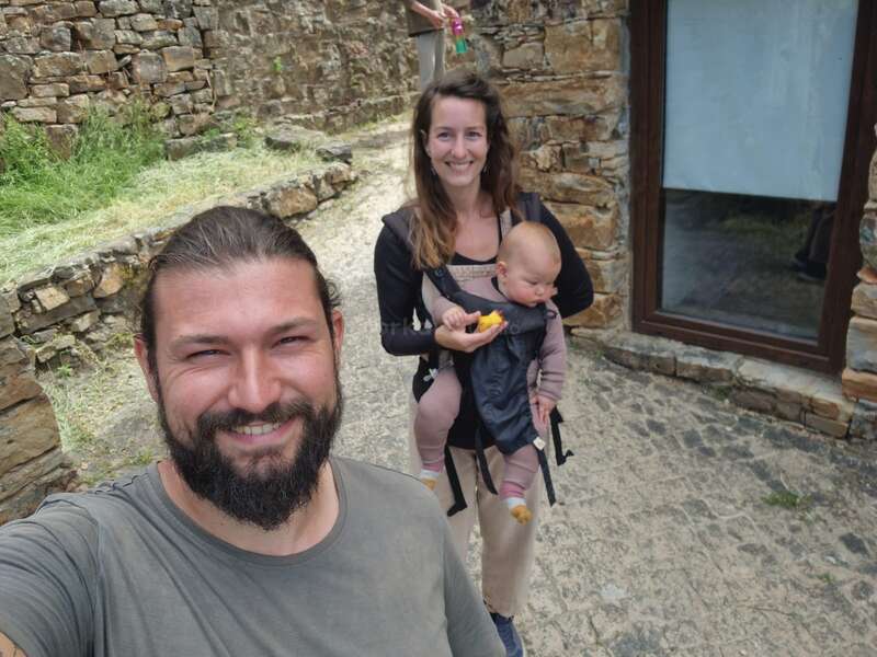 Un homme souriant prend un selfie en plein air, sur un chemin pavé, tandis qu'une femme tenant un bébé dans un porte-bébé se tient debout à côté d'un mur de pierre.