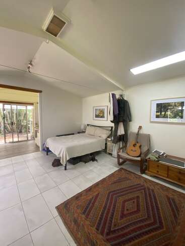 A cozy bedroom with white walls, a single bed, clothes hanging, armchair with guitar, wooden drawers, large patterned rug, and glass doors leading outside.