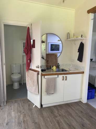 A small kitchen with a round mirror, white cabinets, a sink, and towels. An open door reveals a toilet. Wooden floor and minimal decorations. Cozy space.
