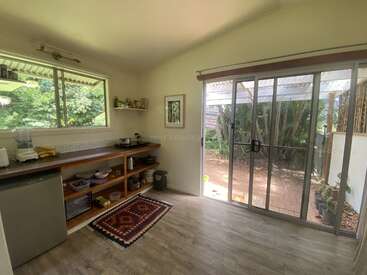 This cozy kitchen features wooden shelves, a small fridge, and a patterned rug. Large sliding glass doors and a window offer abundant natural light and garden views.